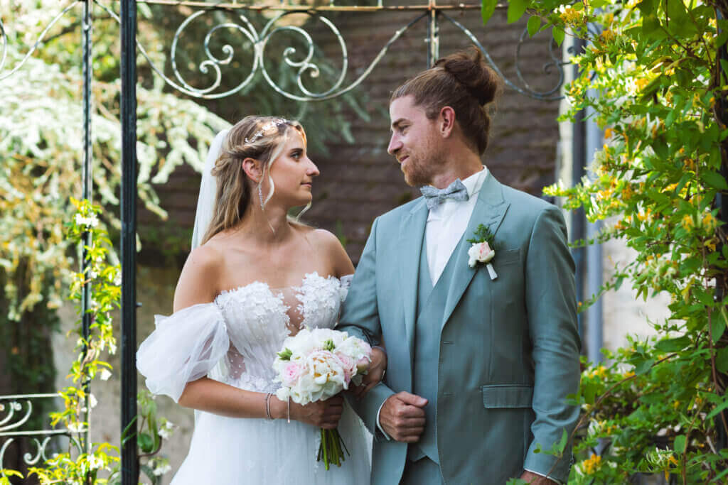 Les mariés se regardent dans un passage végétalisé, la mariée tenant son bouquet et le marié en costume vert clair.
