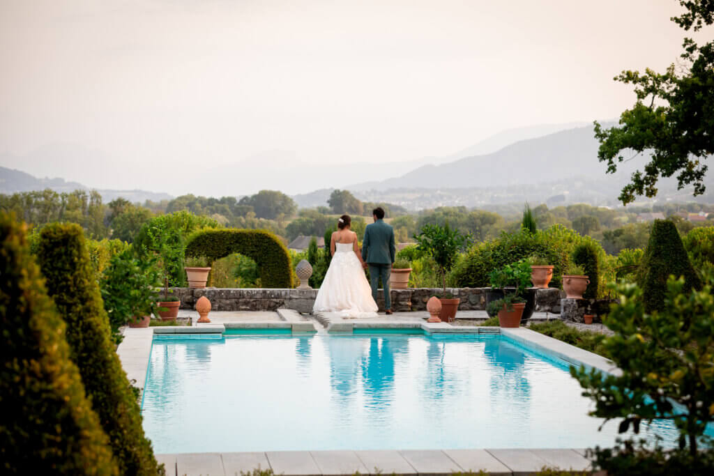 Couple de mariés de dos marchant près d’une piscine avec vue dégagée sur un paysage vallonné.