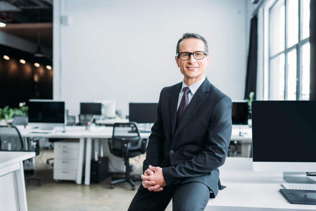 Portrait d'un professionnel en costume souriant dans un open space moderne avec lumière naturelle et postes de travail en arrière-plan