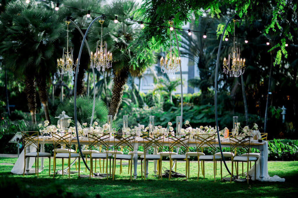 Table de réception de mariage en extérieur décorée de fleurs blanches et chandeliers suspendus dans un jardin verdoyant