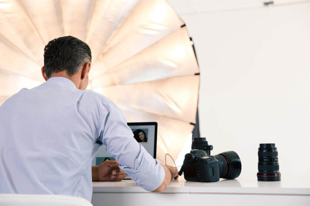 Photographe professionnel vu de dos travaillant sur un ordinateur portable en studio avec boîtier et objectifs posés sur le bureau