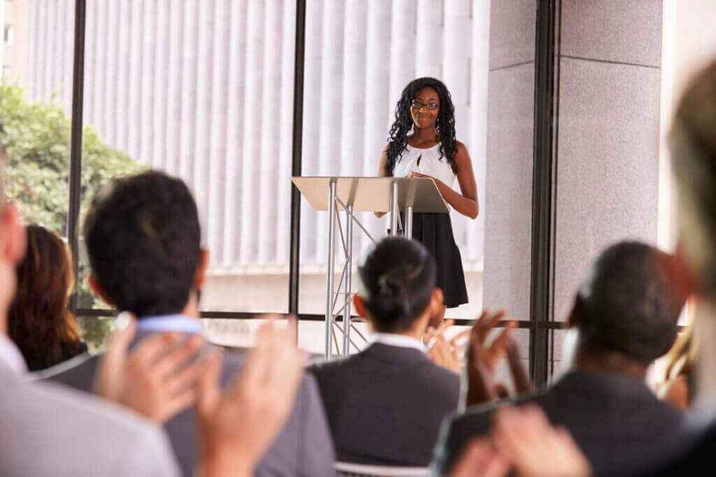 Femme prenant la parole à un pupitre lors d'une conférence d'entreprise avec le public applaudissant au premier plan